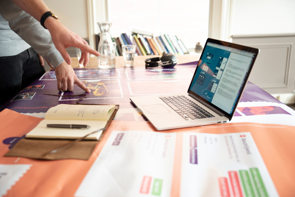 Hand wijst naar laptop op tafel vol design attributen